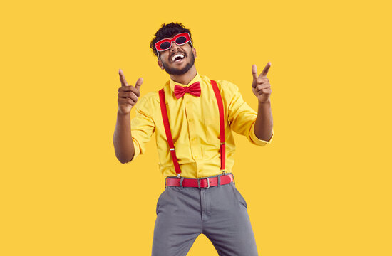 Funny stylish bearded indian guy having fun and dancing isolated on orange background. Cool young ethnic man in shirt with bow tie and pants with suspenders laughs and dances in funny glasses.