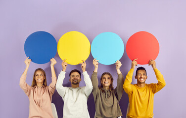 Different happy cheerful positive young multiethnic people sharing their points of view holding round colourful red, blue and yellow mock up speech bubble banners. Studio portrait on purple background