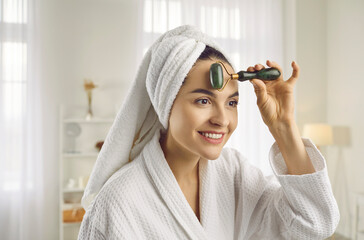 Pretty young woman with bath towel on head massaging face with trendy beauty facial roller to...