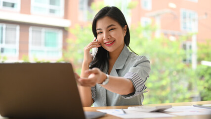 Charming asian businesswoman consulting client on cellphone and using laptop computer at modern office