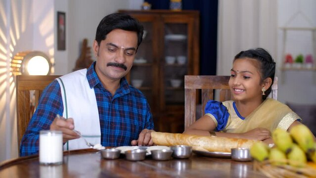 A middle-aged South Indian father feeding his lovely daughter - traditional dress  South Indian family  Single father. An Indian man and his little kid eating food together at the dining table - a ...