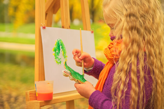 little girl artist with a brush and paints in her hands in autumn in the park draws a landscape with leaves on canvas.
