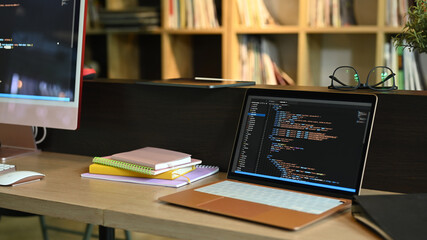 Laptop and computer pc on wooden office desk with programming code on screens