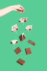 Minimal composition with chocolate brownies covered.with white cream floating against green background. Female hand pouring crumbs on cookies. Tasty dessert concept. Abstract food visual.