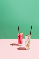 Minimal composition made of two cocktails in highball glasses with straws and sunny day shadow against pastel pink and green background. Bars and summer party concept.