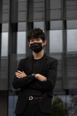 Portrait of an Asian man wearing a black suit and mask standing with arms crossed looking at the camera with a building in the background