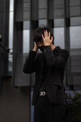 Portrait of an Asian man wearing a black suit and mask feeling stressed by touching his head with both hands with a building in the background