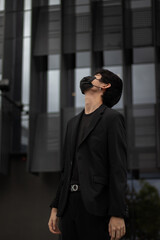 Portrait of an Asian man wearing a black suit and mask closes his eyes and looks up at the sky with a building in the background
