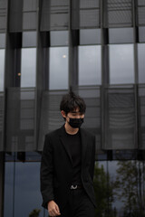 Portrait of a walking Asian man wearing a black suit and mask with a building in the background