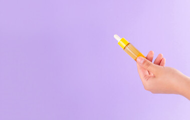 glass yellow bottle with pipette with essential oil in female hand on purple background top view. Aromatic cosmetic product for skin hair care Moisturizing beauty product with vitamin C close up