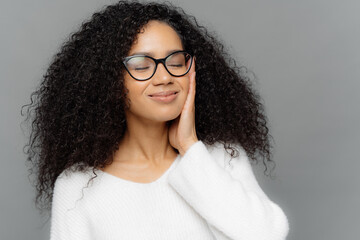 Portrait of satsfied dark skinned woman with curly hair, has eyes closed, touches cheek with hands, imagines something pleasant, wears optical glasses and white jumper, poses indoor. Close up portrait