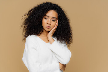 Melancholic sad lonely woman focused down with thoughtful expression, keeps hand on cheek, has calm expression, wears white sweater, has curly hair, isolated on brown studio wall. Depression