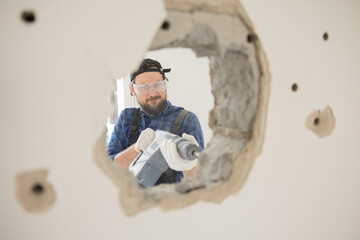 Forging a hole in a wall out of the way with an impact hammer. A construction worker wearing safety goggles in a work suit.