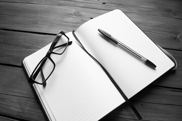 Notepad, pen and glasses. Black and white photo of a notebook with a pen and glasses on a wooden...