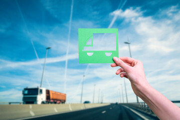 Hand holds green logistics truck symbol against highway - 518759612