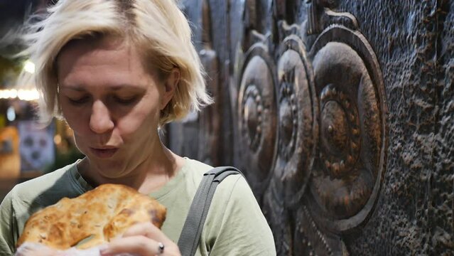 Portrait: An adult young hungry woman bites off a bread roll sprinkled with seeds with her teeth. A woman bought crispy hot bread and eats it outdoors with a good mood and appetite
