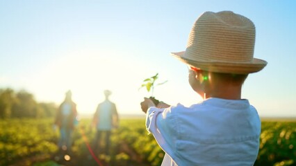 Boy child is holding young tree plant in ground in his palm hands, his parents walking towards him in background. Outdoor nature gardening in field. Care on ecology and environmental protection. - Powered by Adobe