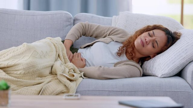 Young woman in pain, suffering from a stomach ache or cramps lying on a sofa sick on her menstrual period at home. African American female feeling ill and holding her belly in discomfort due to ibs