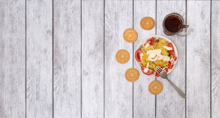 On wooden table are plate with rice porridge and tomato slices, fork, glass of tea and cookies. View from above.