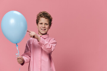 cute little school-age boy stands with a blue balloon in his hand and points at it with his finger...