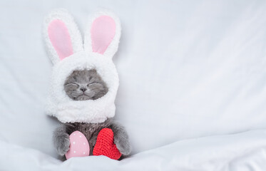 Funny kitten wearing easter rabbits ears sleeps with red heart and painted egg on a bed under warm white blanket at home. Top down view. Empty space for text