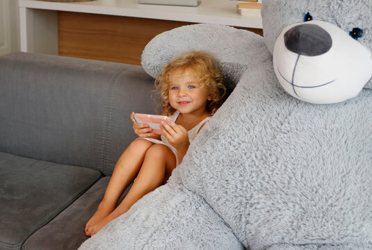 Cute happy little girl with smartphone sitting on sofa with big toy bear.