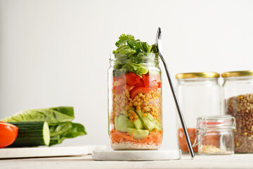 Trendy Fresh detox summer salad layered in a glass mason jar: yoghurt dressing, carrots, cucumber, yellow lentils, red bell pepper and green leaves and herbs, long silver spoon, on white background