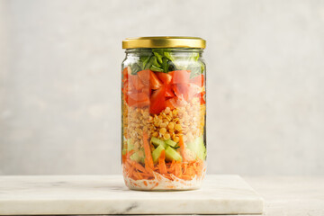 Trendy Fresh detox summer salad layered in a glass mason jar: yoghurt dressing, carrots, cucumber, yellow lentils, red bell pepper, green leaves and herbs, long silver spoon, on white background