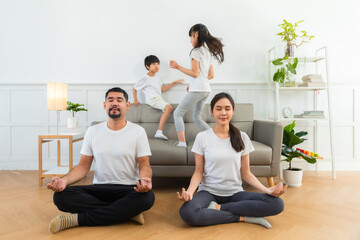 Happy father and mother with closed eyes meditating in lotus pose on floor harmony while excited children jumping on sofa .