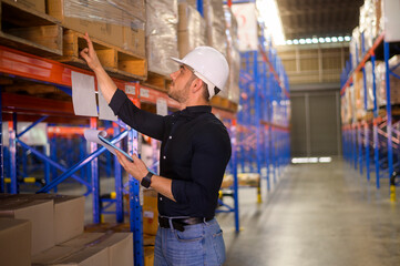 Young worker wearing helmet checking inventory and counting product on shelf in modern warehouse.