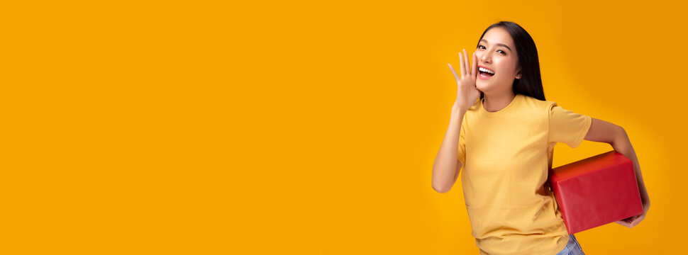 Cheerful young asian woman holding red gift box and looking at camera Standing over yellow background Beautiful Asia girl happy smile hold new year or birthday gift box present Telling something