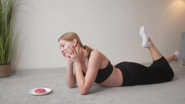 Sweet addiction. Healthy nutrition. Weight loss motivation. Relaxed athletic woman in activewear enjoying doughnut flavour lying on floor.