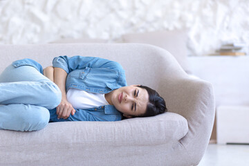 Young beautiful woman alone at home, lying on the sofa, close-up photo, upset, having a severe stomach ache