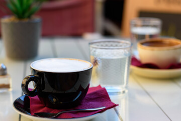 Cup of cappuccino coffee in black cup, spoon on saucer, cup of greek coffee with glass of water on a wooden white table at the cafe.
