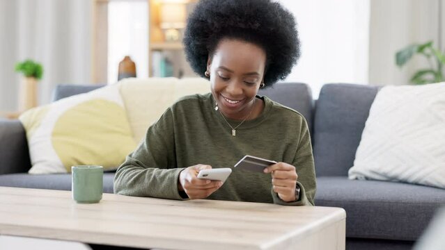 Young black woman shopping online using her phone and bank credit card in a bright living room. Banking and managing assets and finance using convenient wireless technology from the comfort of home