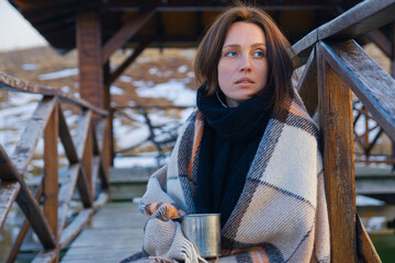 A beautiful girl wrapped in a plaid walks along the bridge over the pond with a metal mug of tea on...