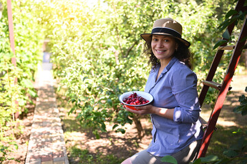 Beautiful woman, eco farmer smiling toothy smile, looking at camera, holding a bowl with freshly picked ripe cherries