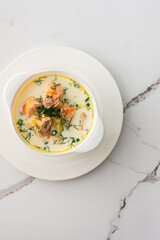 Norwegian red salmon fish soup with vegetables, herbs and cream, top view of a bowl of soup on a white marble background