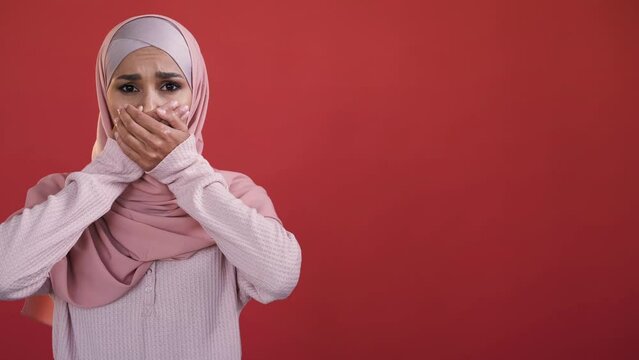 Female rights. Woman discrimination. Gender equality. Upset scared speechless lady in hijab covering mouth with hand isolated on red empty space background.
