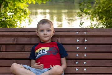 adorable little boy sitting on a park bench on the background of the water surface. close-up image