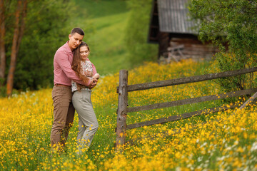 Fototapeta premium Young loving couple gently hugging on the wood house background, a lot of yellow flowers are around on the field. Enjoying time together. 