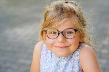 Portrait of a cute preschool girl with eye glasses outdoors. Happy funny child wearing blue glasses, loooking at the camera. Sunny summer day in the city.