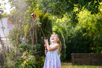 Happy little girl playing with a garden hose on hot and sunny summer evening. Preschool child helping and having fun with watering trees and plants in domestic garden
