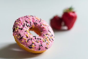 A photo of a donut with pink icing, a small part of which is in focus and a strawberry in the background, very out of focus. All objects on a light background