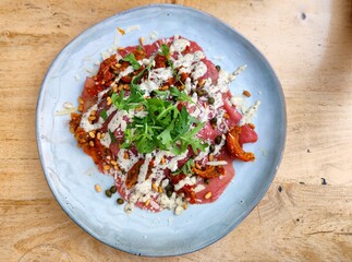 Tenderloin carpaccio with lettuce, cucumber, sun-dried tomatoes, capers, pine nuts, parmesan, arugula and pesto truffle dressing on a board on a restaurant table. There are no people or trademarks.