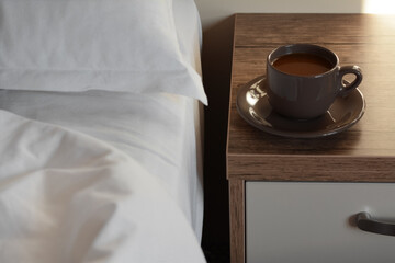 Fototapeta na wymiar Cup of morning coffee on wooden night stand near bed indoors