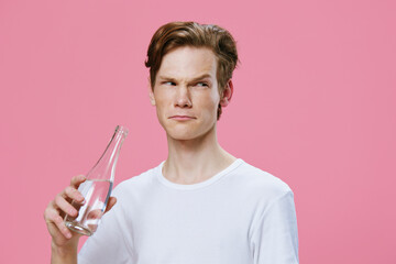 handsome attractive guy with red hair in a white T-shirt thoughtfully twisted his face holding a glass bottle of water in his hand standing on a pink background with empty space for advertising text