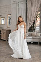Fototapeta premium The beautiful woman posing in a wedding dress. Young beautiful happy bride wearing white wedding dress and posing in bright empty interior. Wedding fashion. Expensive Lace bust.