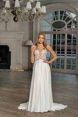 Fototapeta premium The beautiful woman posing in a wedding dress. Young beautiful happy bride wearing white wedding dress and posing in bright empty interior. Wedding fashion. Expensive Lace bust.
