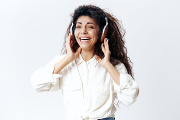 Smiling Curly Beautiful Latin Woman Listening Cool Playlist In Headphones Singing Look At Camera Isolated At White Studio Wall Background. Music Devices DJ Technologies Huge Sale Concept. Copy space